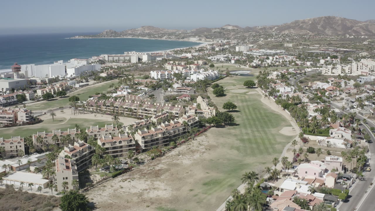 San Jose del Cabo aerial frame with a long green fairway, a curving road at the right edge, clustered homes, blue ocean, and hazy hills, with no visible people.