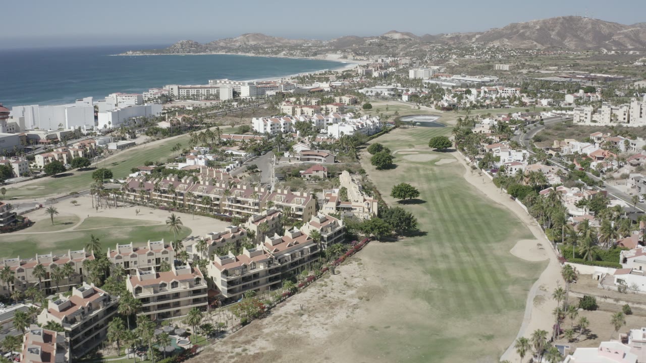Aerial view of coastal homes, golf course, beach, sea, and hills.