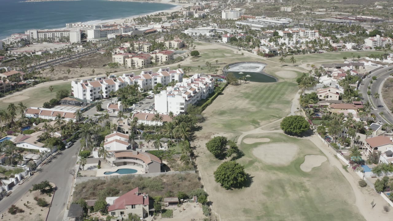 Aerial view in San Jose del Cabo with the golf course centered, white apartment-style buildings to the left, curved roads to the right, coastline in the distance, and no visible people.