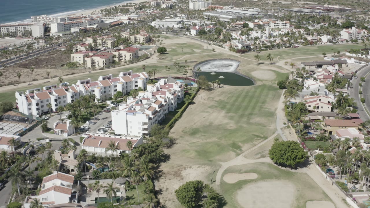 Aerial view in San Jose del Cabo with white multi-story housing in the foreground, golf course greens and pond in the middle, and ocean beyond, with no visible people.