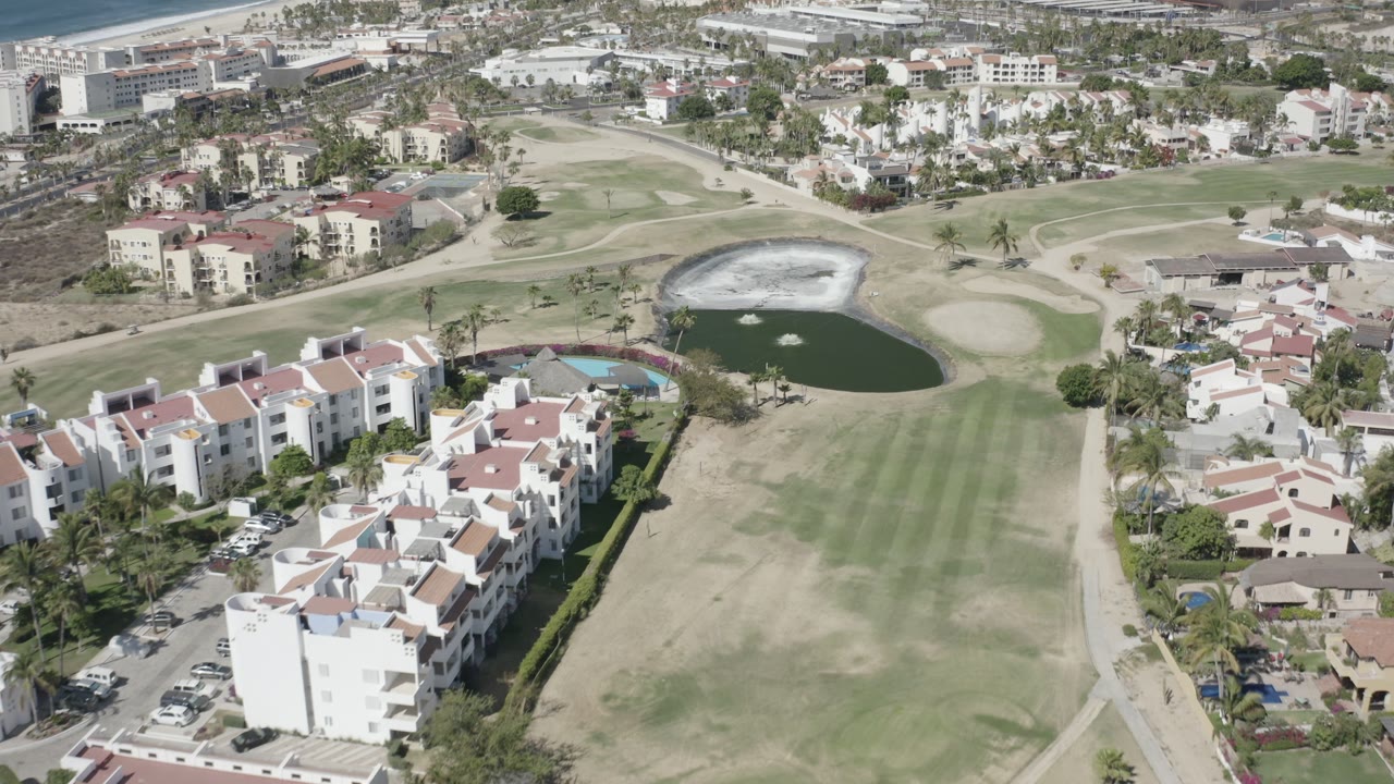 High aerial view in San Jose del Cabo, Mexico, showing white residential buildings, a fairway, sand traps, a pond with fountains, and no visible people.