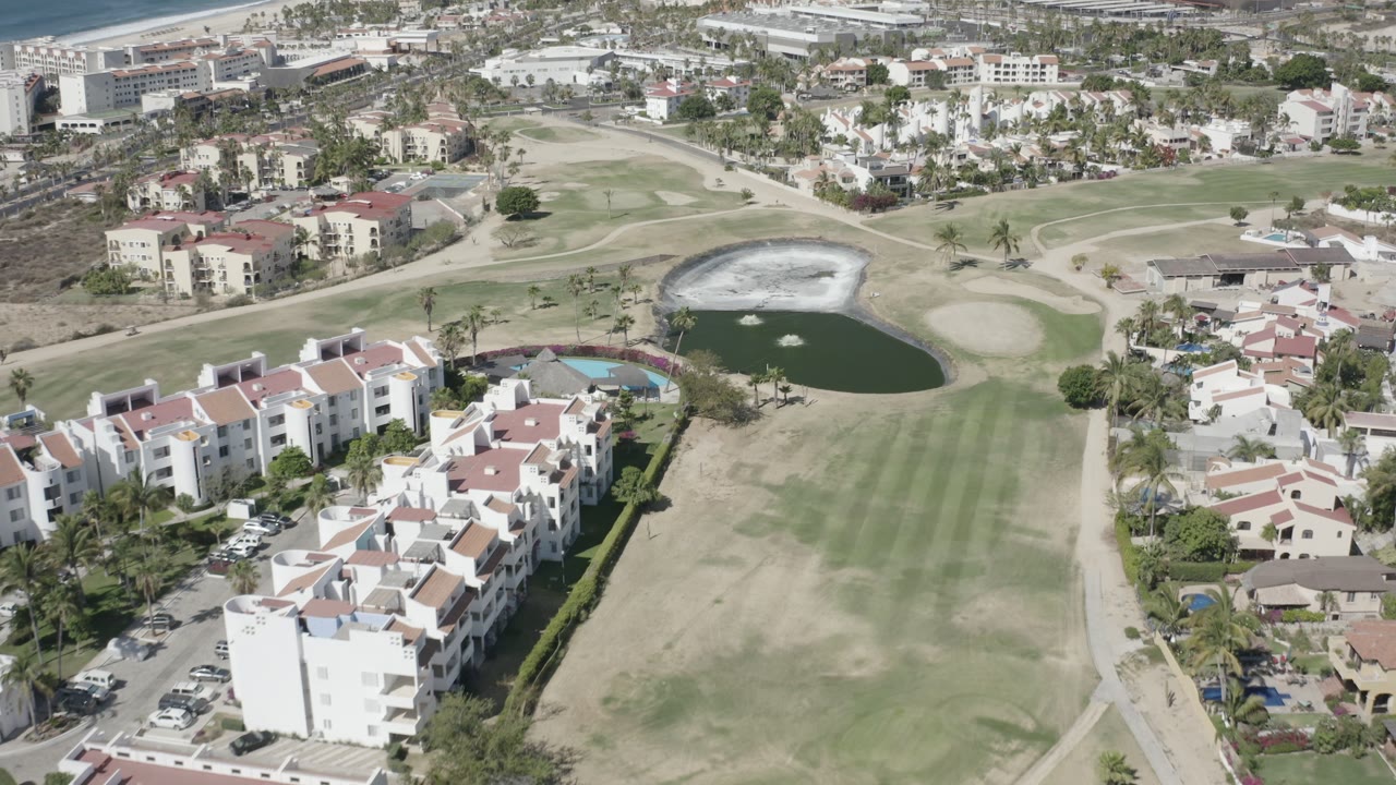 Elevated drone frame in San Jose del Cabo, Mexico, with a central fairway, dark pond, pale bunkers, red-roof homes, and no visible people.