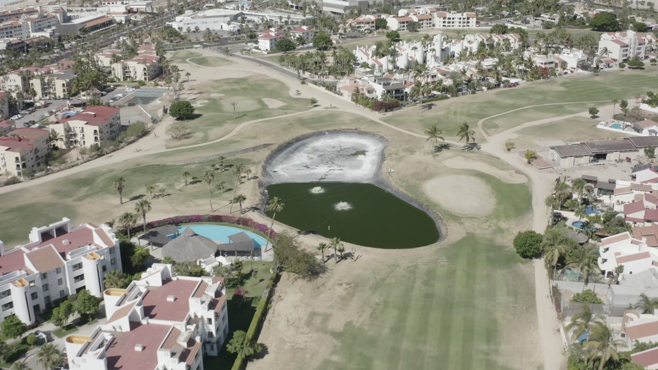 Overhead view of golf-course grass, pond, pool, and residential buildings.