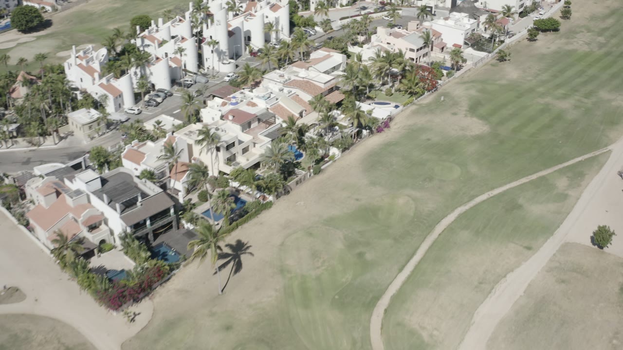 Aerial frame from San Jose Del Cabo with the fairway filling the right side, clustered homes and palms on the left, and no clearly discernible faces.