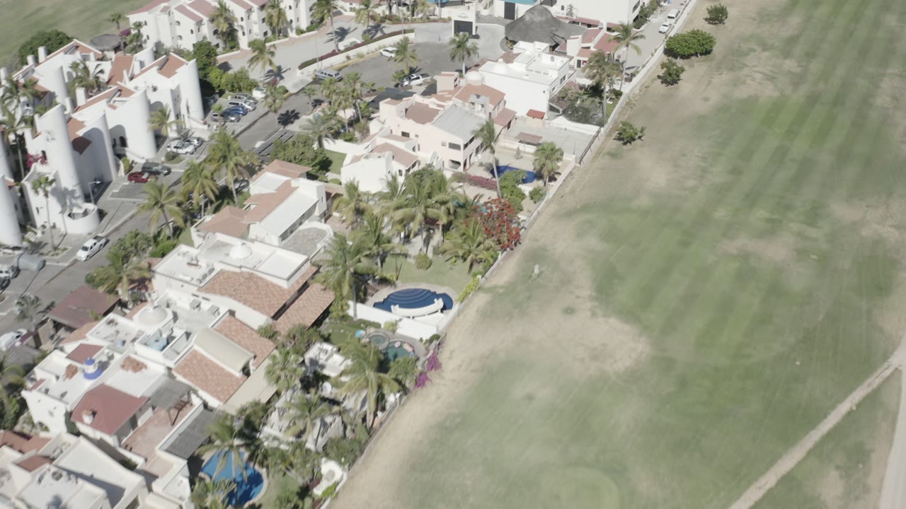 Aerial view in San Jose del Cabo with the road and parked vehicles near villas on the left and the grass clearing extending across the right, with no visible people.