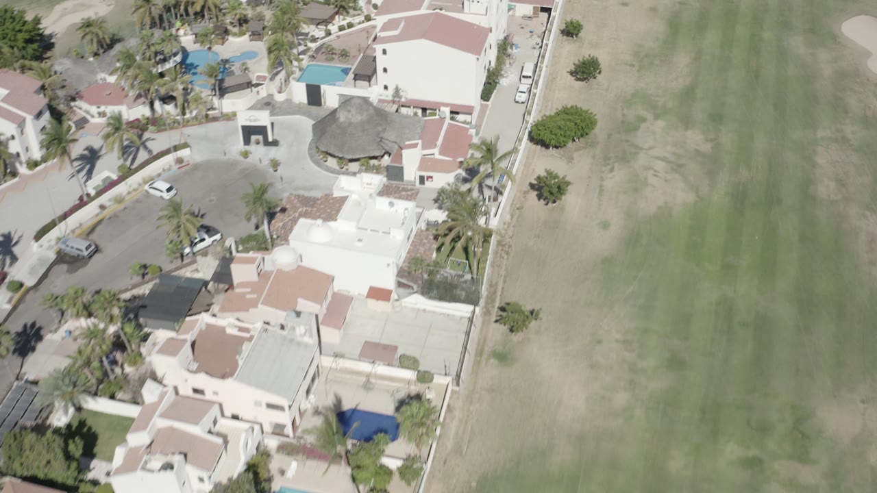 Overhead aerial frame in San Jose Del Cabo, Mexico showing white homes with red roofs beside a broad green field, with palm trees and no visible people.