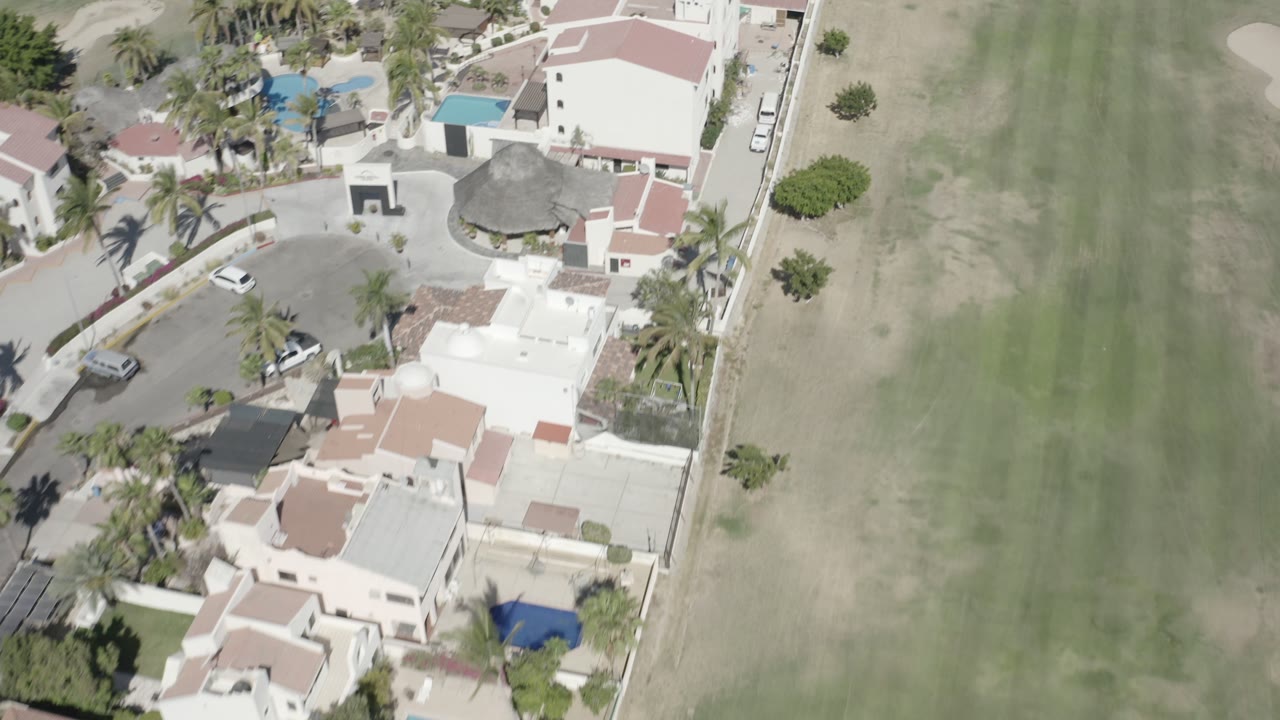 High overhead view in San Jose Del Cabo, Mexico with villas, blue pools, palm landscaping, parked cars, and the green field taking up the right side, with no visible people.
