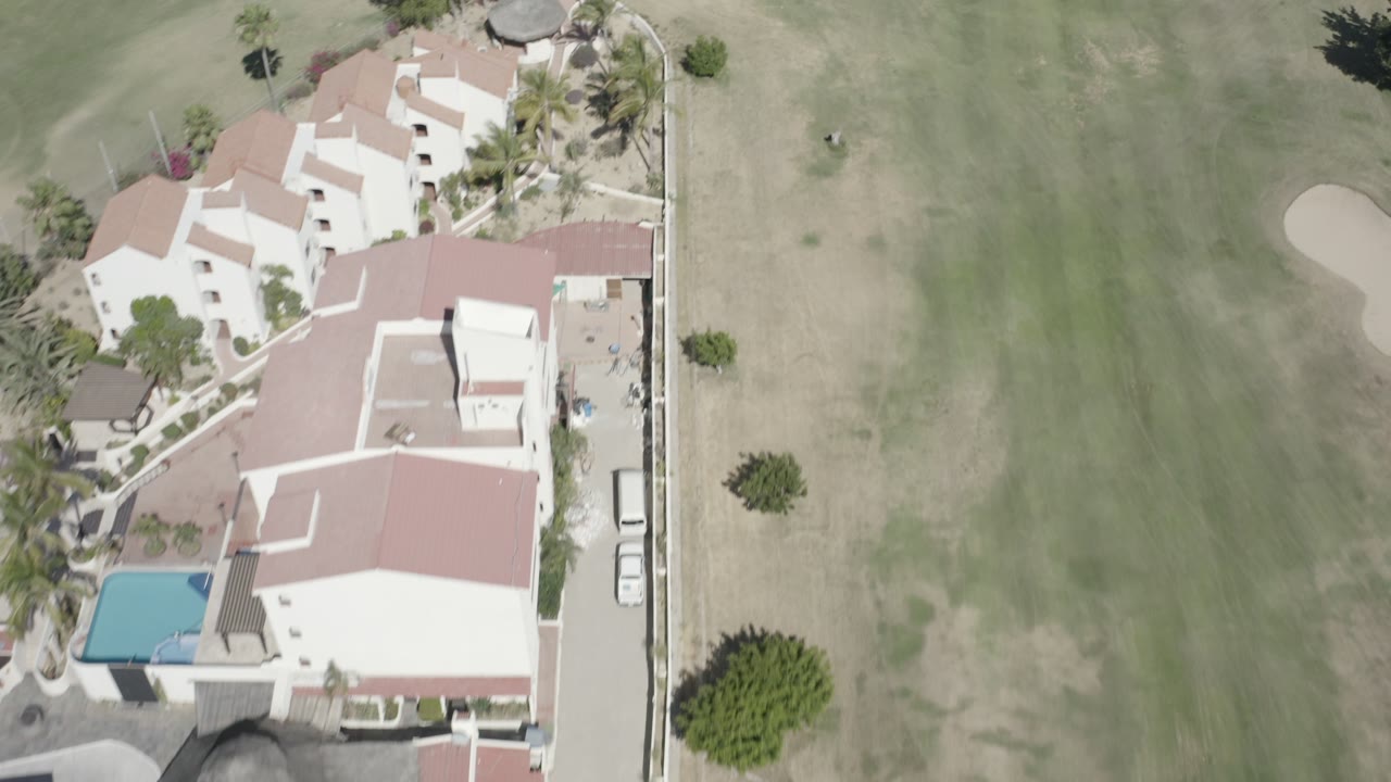 San Jose del Cabo aerial frame with the housing cluster and pool near the left edge, open fairway grass to the right, parked vehicles, and no visible people.