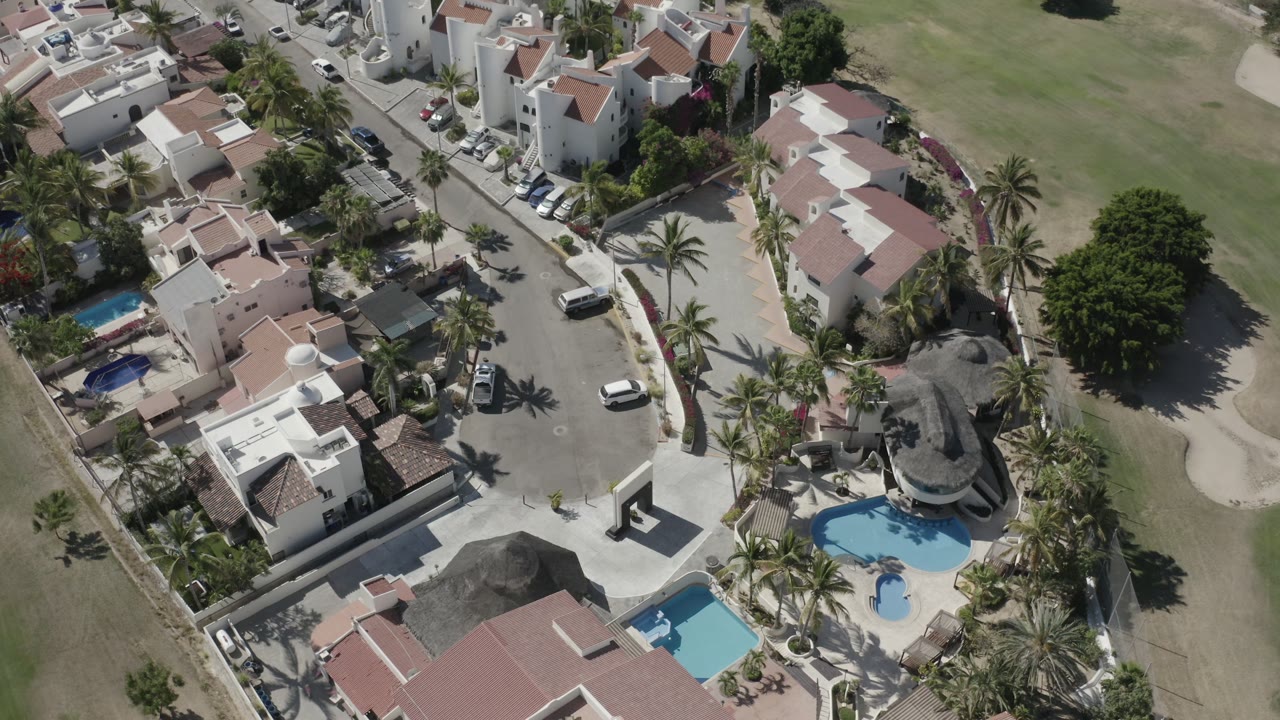 Overhead frame in San Jose del Cabo showing white residential buildings, small private pools, palm-lined streets, parked cars, and a large open grass area, with no visible people.