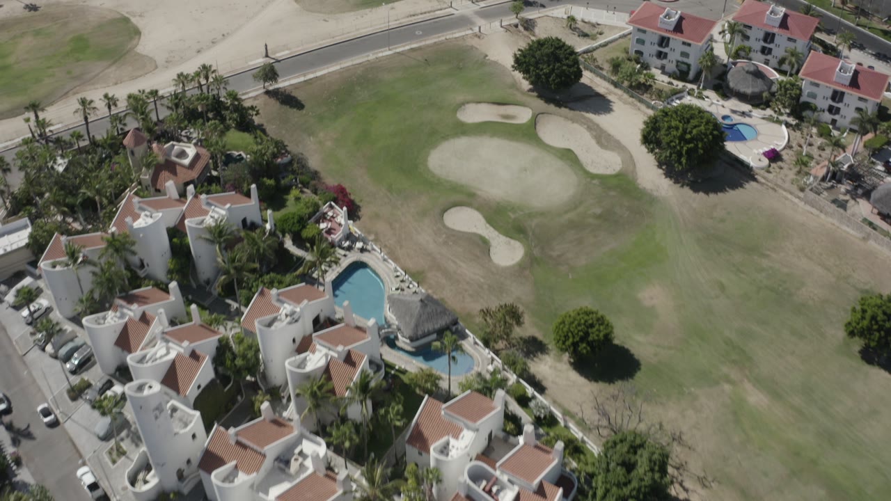 Aerial view over San Jose del Cabo homes with red tile roofs, blue pools, a green golf fairway, pale bunkers, and no visible people.