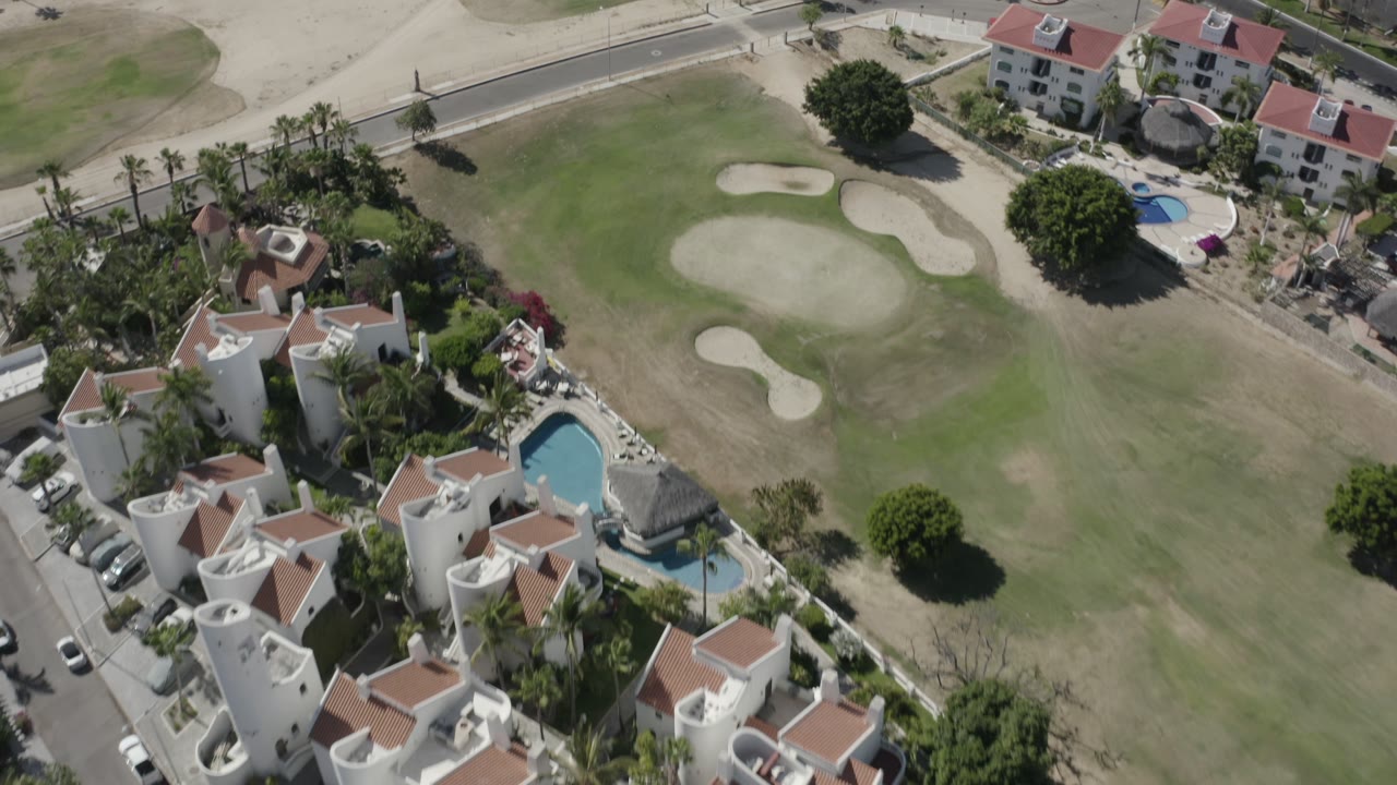 Drone view in San Jose del Cabo with clustered white residences at left, pools below, fairway grass and sand bunkers across the center, and no visible people.