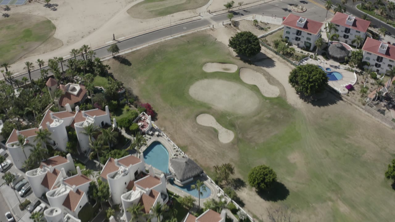 Overhead angle on golf greens, sand traps, villas, pools, and nearby buildings.