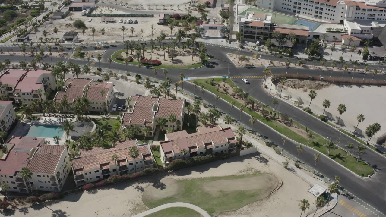 Aerial frame over San Jose Del Cabo showing intersecting roads, red-roofed low-rise buildings, palm trees, sandy ground, and a small pool area with no visible people.
