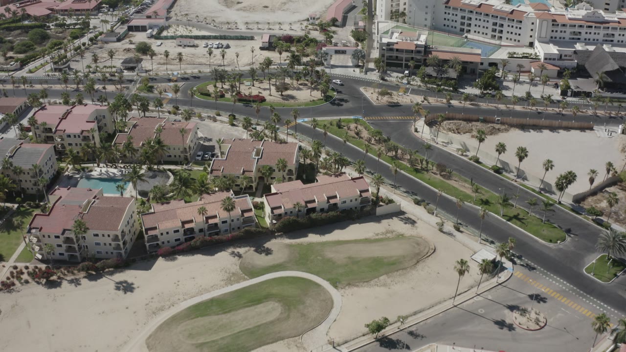 Aerial frame over San Jose del Cabo resort buildings with red roofs, crossing roads, palm trees, sandy lots, and larger hotel blocks in the background.