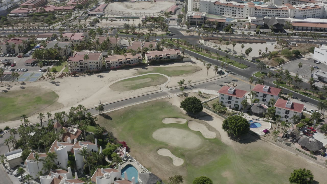 A high aerial frame over San Jose del Cabo shows a golf fairway, sand bunkers, red-roofed homes, roads, palm trees, and no visible people.
