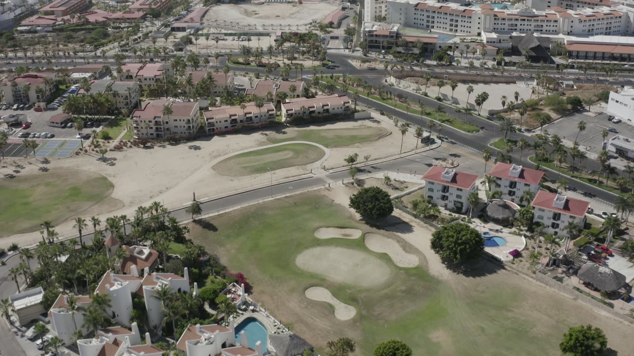 The second frame in San Jose del Cabo holds above the fairway and bunkers, with a road, palms, clustered homes, and no visible people.