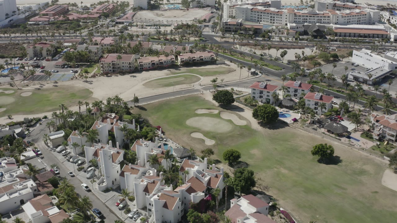 Drone-height view in San Jose del Cabo showing white residential buildings in the foreground, broad fairway grass, pale bunkers, roads, and no visible people.