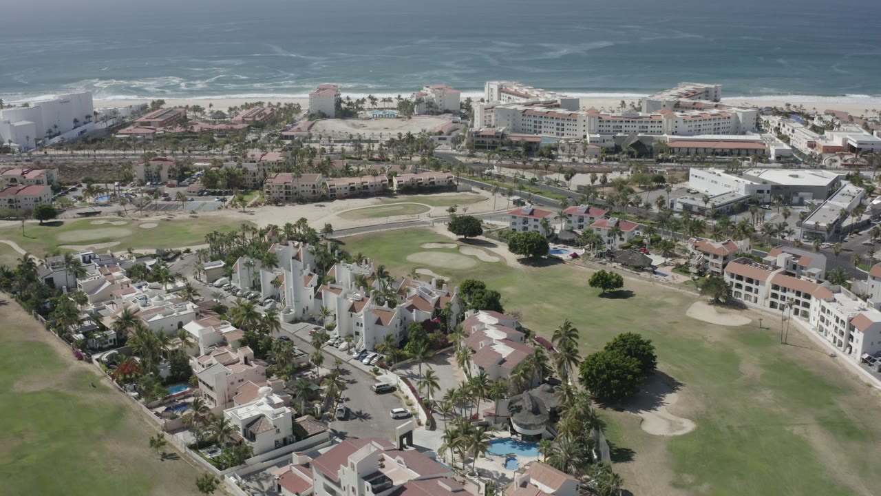Opening aerial frame over San Jose Del Cabo, Mexico, showing low-rise homes, green lawns, palm trees, beachfront buildings, surf, ocean, and no visible people.