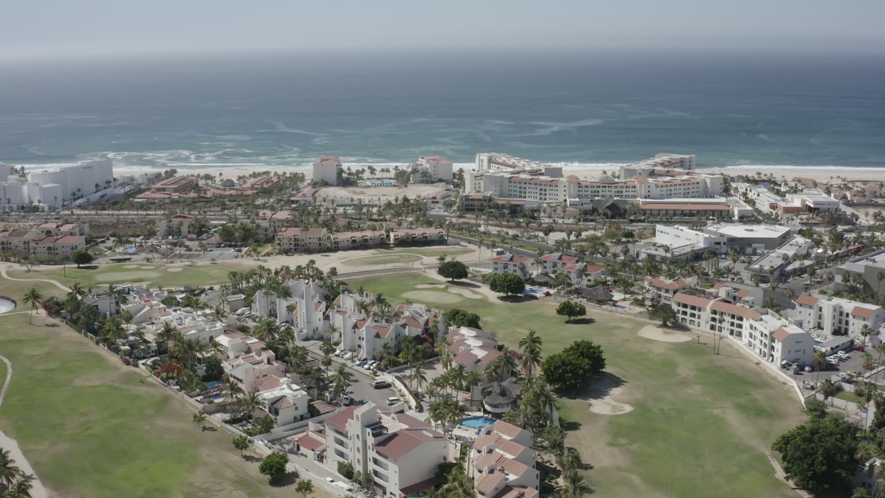 High aerial frame in San Jose del Cabo showing resort-scale buildings near the beach, residential blocks, fairway grass, blue ocean, hazy sky, and no visible people.