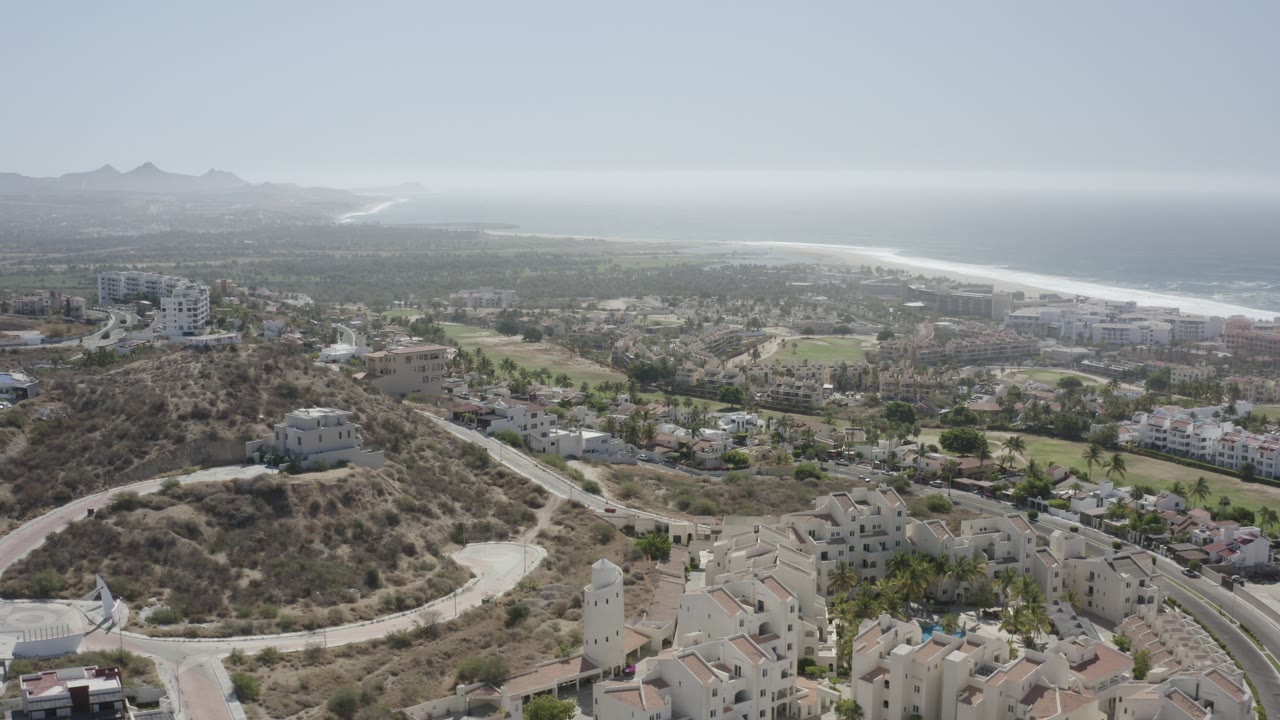 Wide aerial view over hillside buildings, roads, green areas, coastline, ocean, and distant mountains.