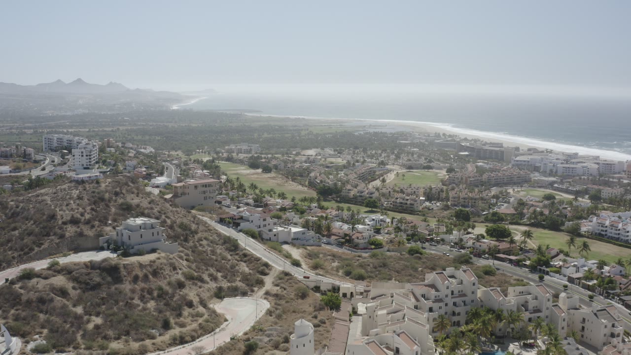 Aerial view over dry hills, roads, buildings, coastline, and distant mountains.