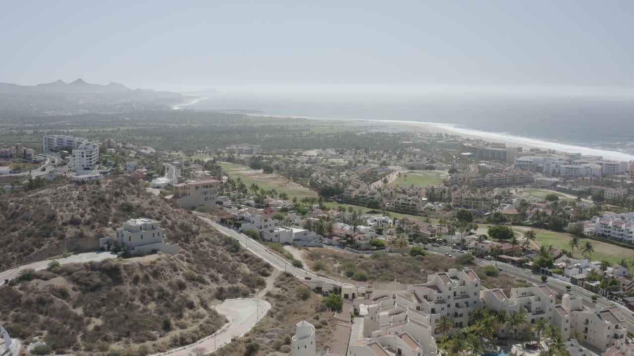 Aerial view in San Jose del Cabo, Mexico, showing a rounded dry hill with pale buildings nearby, a road crossing the lower frame, green open areas, ocean haze, and no visible people.
