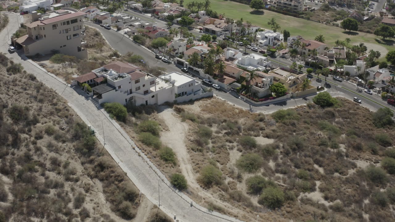 High drone-style view in San Jose del Cabo showing white private houses near a curving road, dry lots, scattered palms, parked cars, and no visible people.