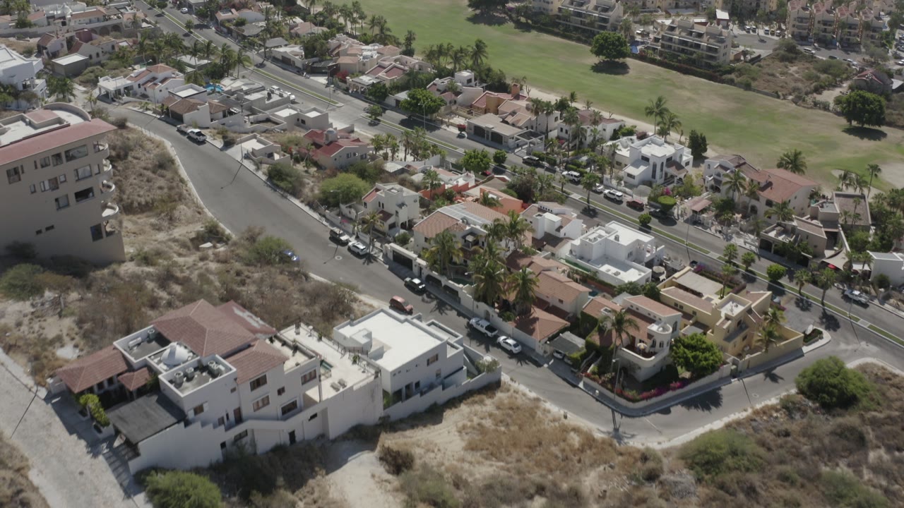 Aerial view of houses, curved roads, parked cars, palms, dry lots, and a green open area.