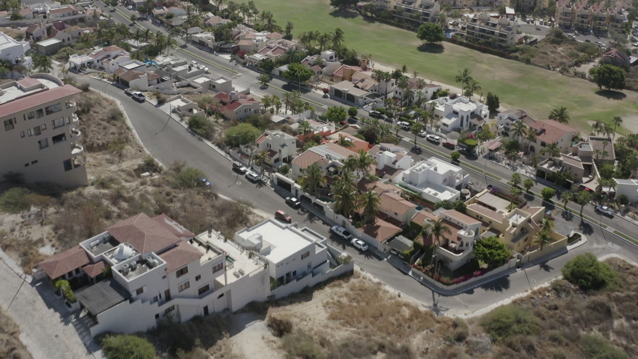 Drone frame over San Jose del Cabo residential streets, showing white homes, parked cars along the curb, palms, dry lots, and no visible people.