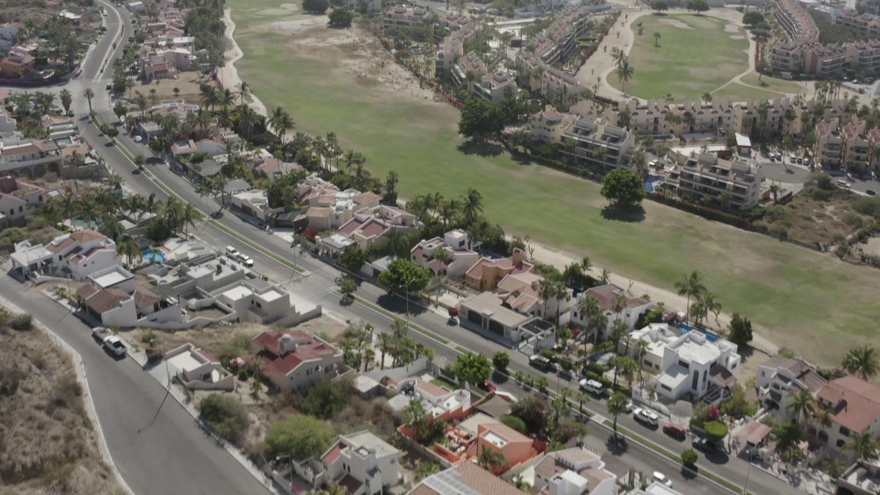 Aerial view in San Jose del Cabo with houses and palm trees in the lower frame, a road crossing diagonally, and broad green fairways beyond, with no visible people.