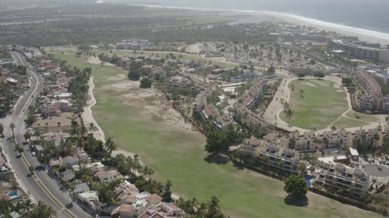 Aerial frame over San Jose del Cabo with fairways in the foreground, clustered homes and palms in the middle, larger shoreline buildings beyond, and no visible people.