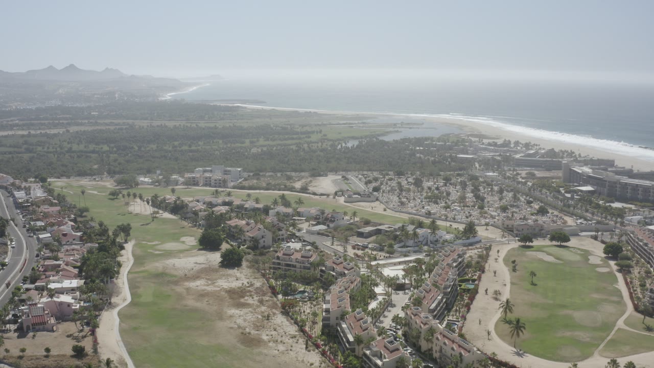 Aerial view of golf course, coastal buildings, beach, sea, and distant hills.