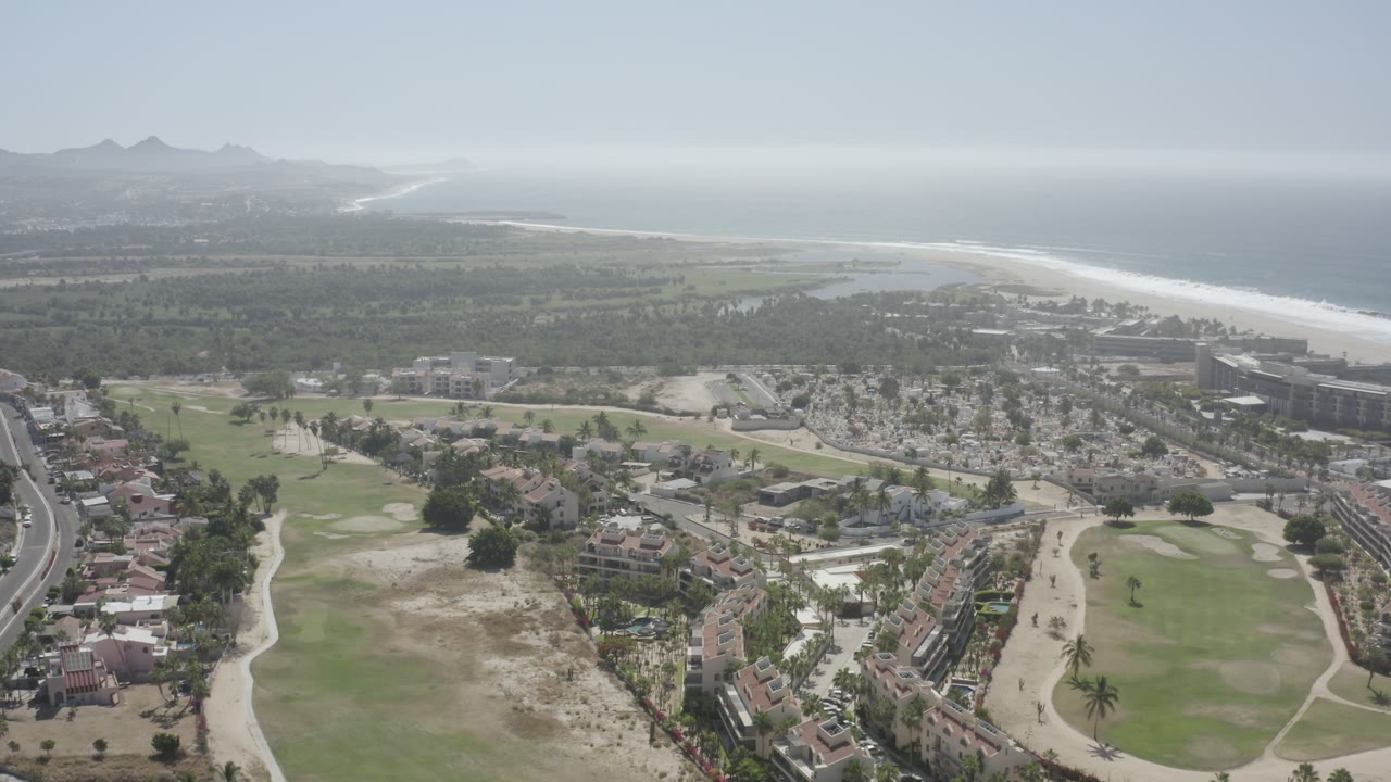 Aerial view in San Jose del Cabo showing more of the curved road at right, golf course greens below, low housing and resort blocks near the shoreline, hazy ocean, and no visible people.