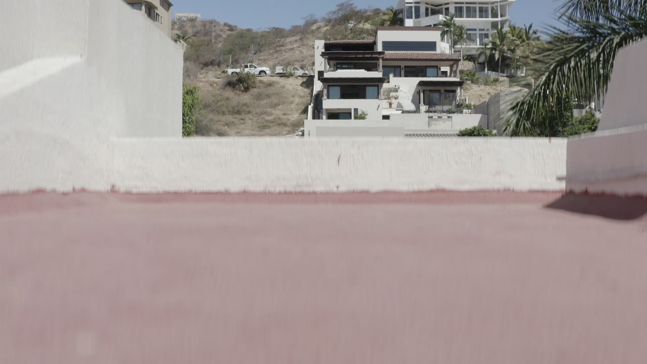 A second early frame in San Jose Del Cabo, Mexico, keeps the white wall and pink roof in the foreground while hillside houses and sparse vegetation remain visible, with no visible people.