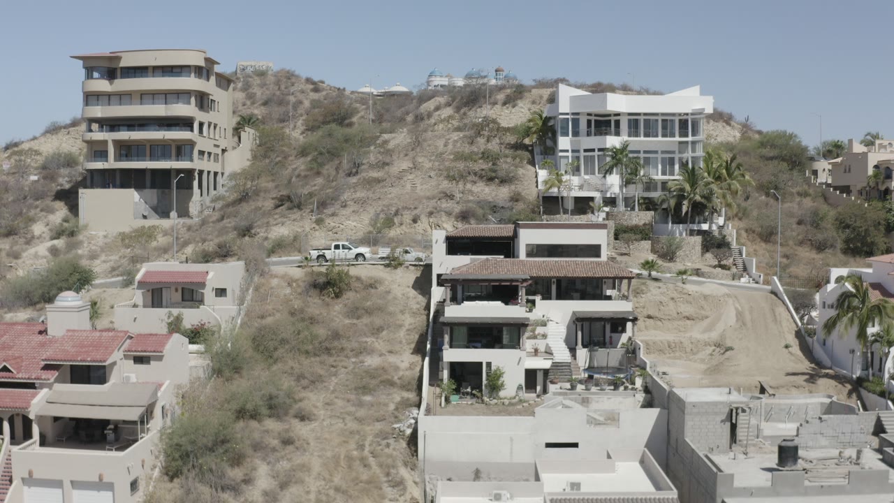 San Jose Del Cabo hillside frame with a multi-story tan house, a white modern house, dry brush, a white pickup, and no clearly visible people.