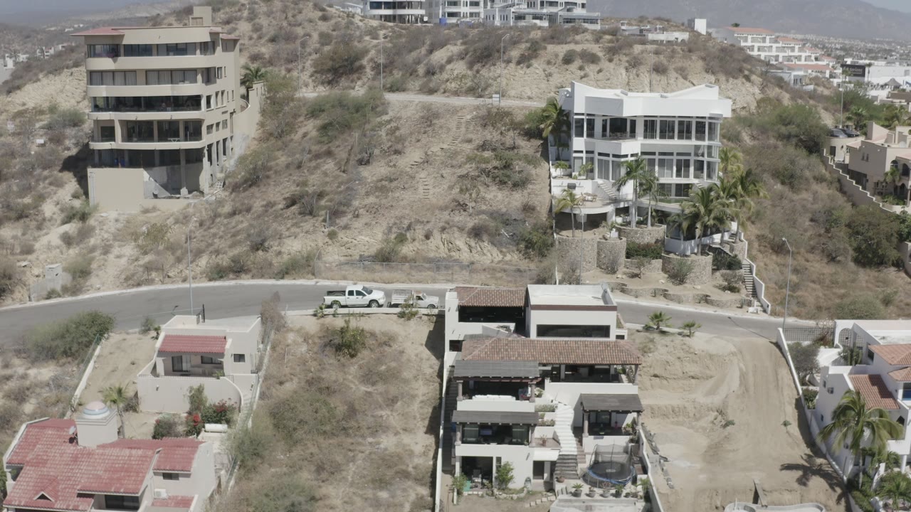 San Jose del Cabo hillside view with a white multi-level house, a darker modern building, dry open lots, palm trees, parked vehicles, and no visible people.