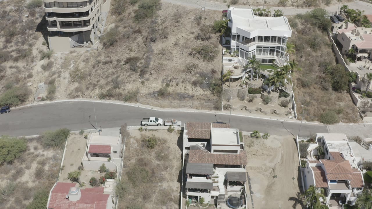 High aerial frame over San Jose del Cabo showing a curved paved road, dry roadside lots, pale homes, parked vehicles, scattered trees, and no visible people.