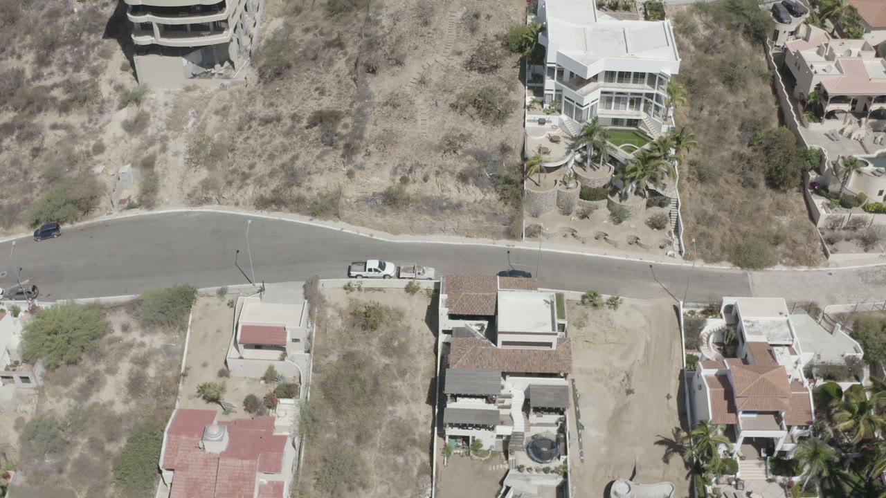 Overhead drone frame in San Jose del Cabo showing a curved paved road between dry vacant land, walled homes, parked vehicles, and no visible people.