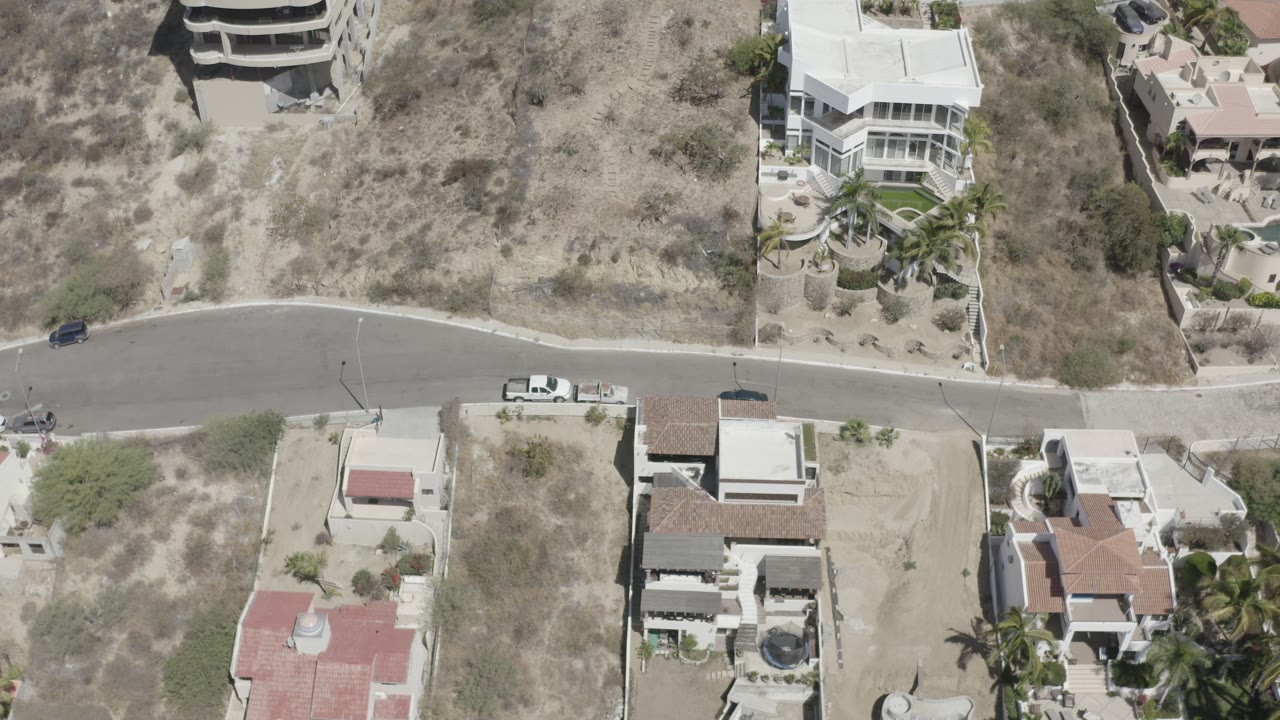 High overhead view in San Jose del Cabo with the empty scrub lot dominating the upper frame, a road through the center, private villas below, and no visible people.
