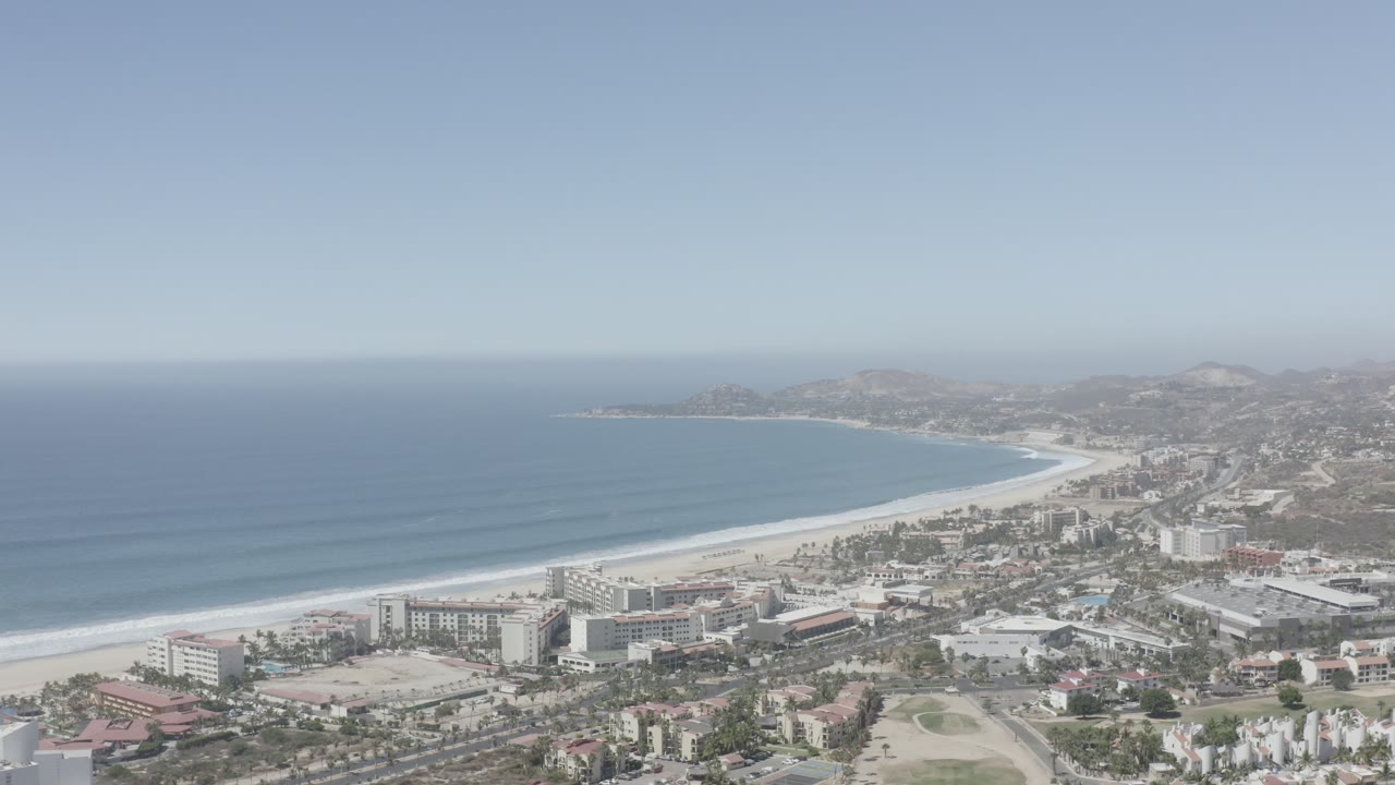 Opening aerial frame over San Jose del Cabo, Mexico, with blue ocean on the left, a pale curved beach, beachfront buildings, inland roads, hazy hills, and no visible people.