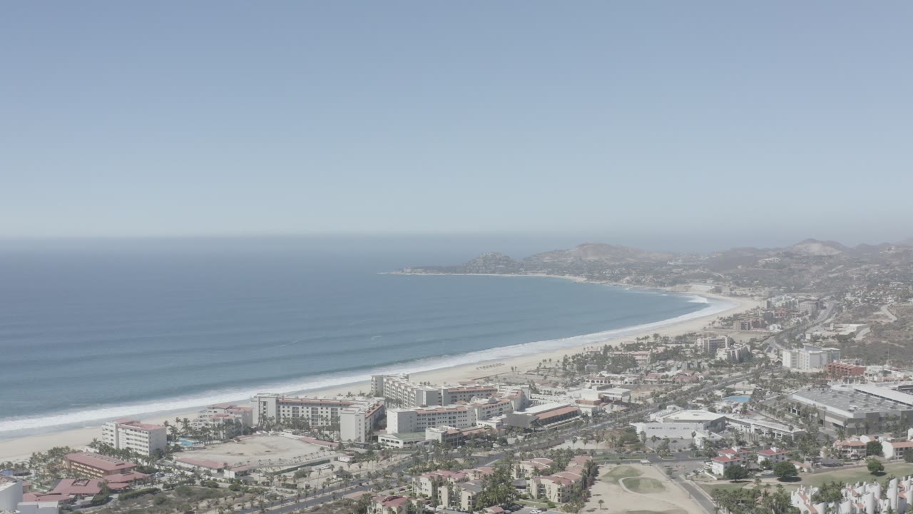 A high view of San Jose del Cabo shows the ocean occupying the left half of frame, a bright beach band, low and mid-rise buildings near shore, distant hills, and no visible people.