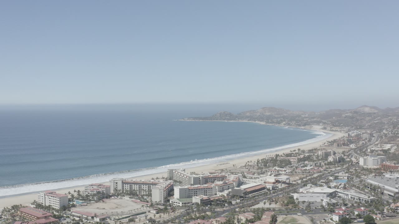 High aerial view of a curved beach, ocean, coastal buildings, and hazy hills.