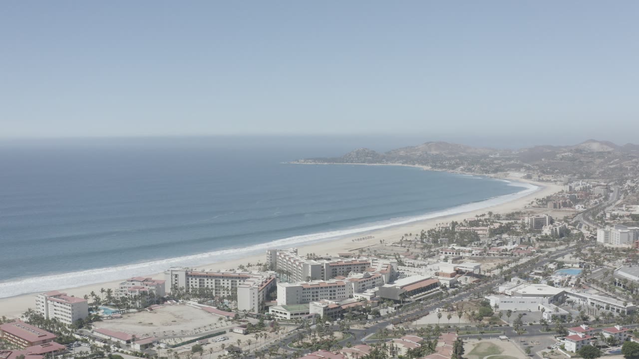 Aerial view of a curved beach, blue ocean, hotels, and hazy hills.