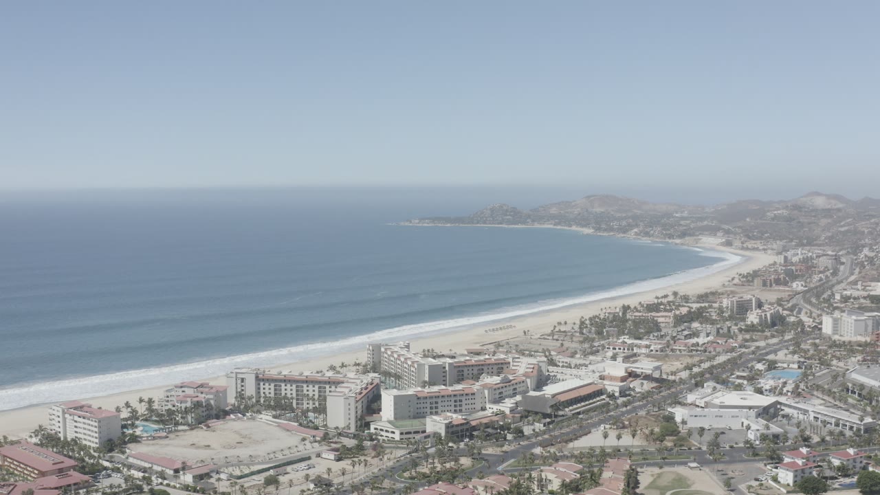 Wide San Jose del Cabo aerial with parallel wave bands offshore, pale sand along the bay, hotel blocks in the foreground, and no visible people.