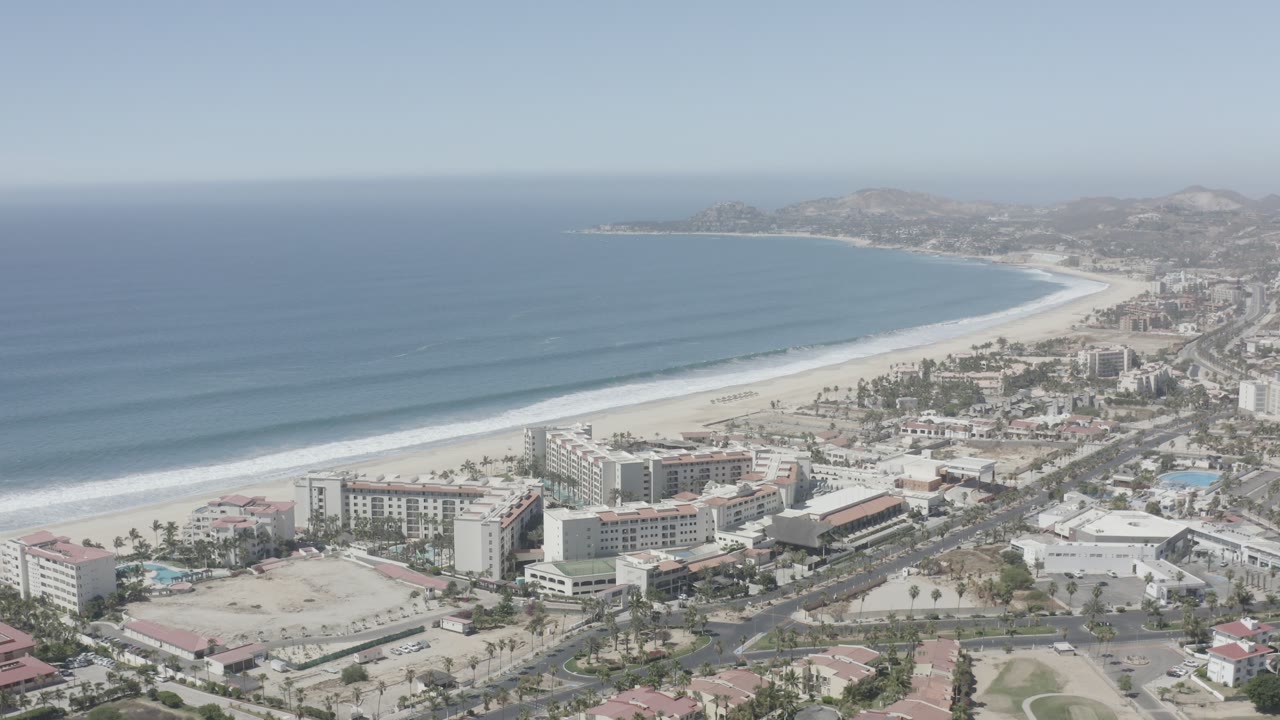 Aerial frame over San Jose Del Cabo coastline with long wave bands approaching the beach, hotels or large coastal buildings, roads, and dry hills in the distance, with no visible people.