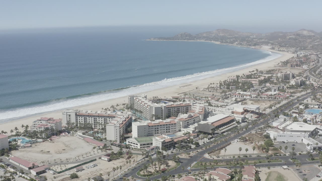 Wide aerial frame of San Jose Del Cabo coastline with a curving beach, parallel surf bands, resort-like buildings near the shore, roads in the lower right, and no visible people.