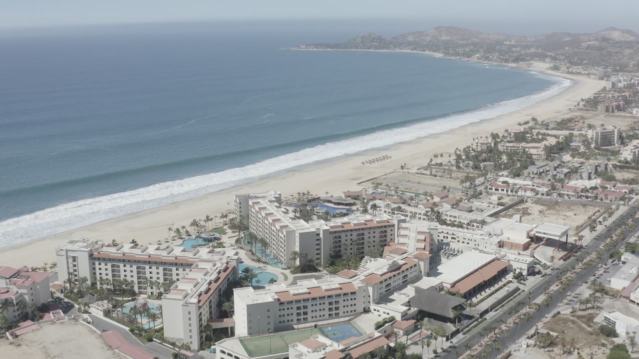 A wide aerial frame over San Jose del Cabo shows blue ocean, white surf, pale beach, resort buildings, pools, a coastal road, distant shoreline, and no visible people.