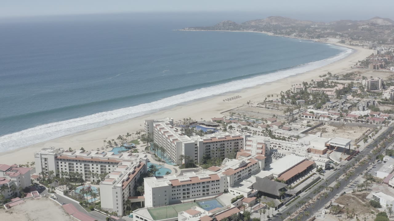 An elevated San Jose del Cabo coastline frame keeps the ocean on the left, surf crossing the beach, resort buildings and pools below, roads to the right, and no visible people.