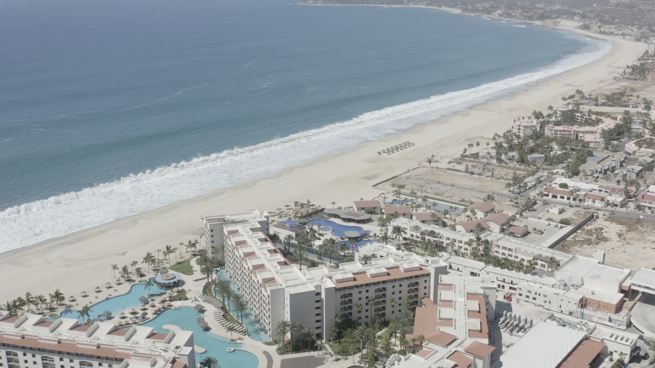 Aerial frame in San Jose del Cabo looking across white hotel blocks and blue pools toward beach umbrellas, rolling surf, and open water, with only distant people visible.