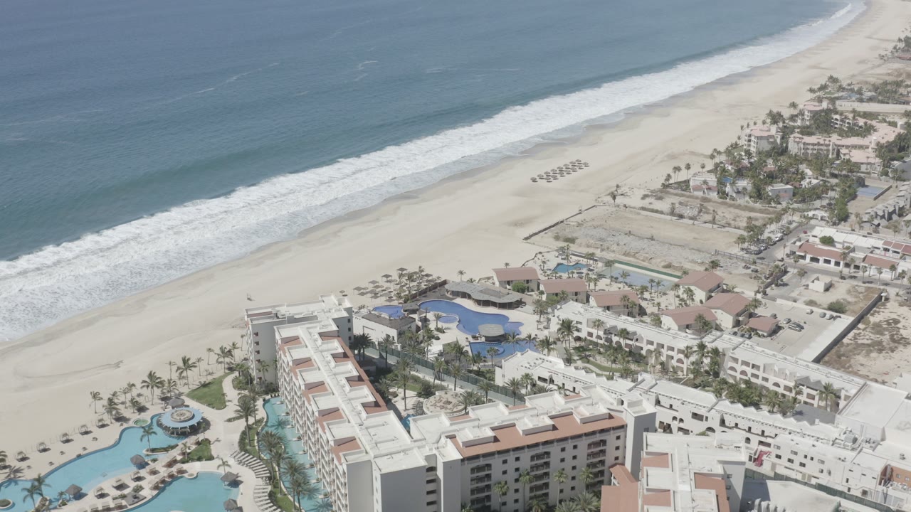 Aerial view over San Jose Del Cabo resort buildings, pools, pale sand, and blue surf at the start of the clip, with no visible people.